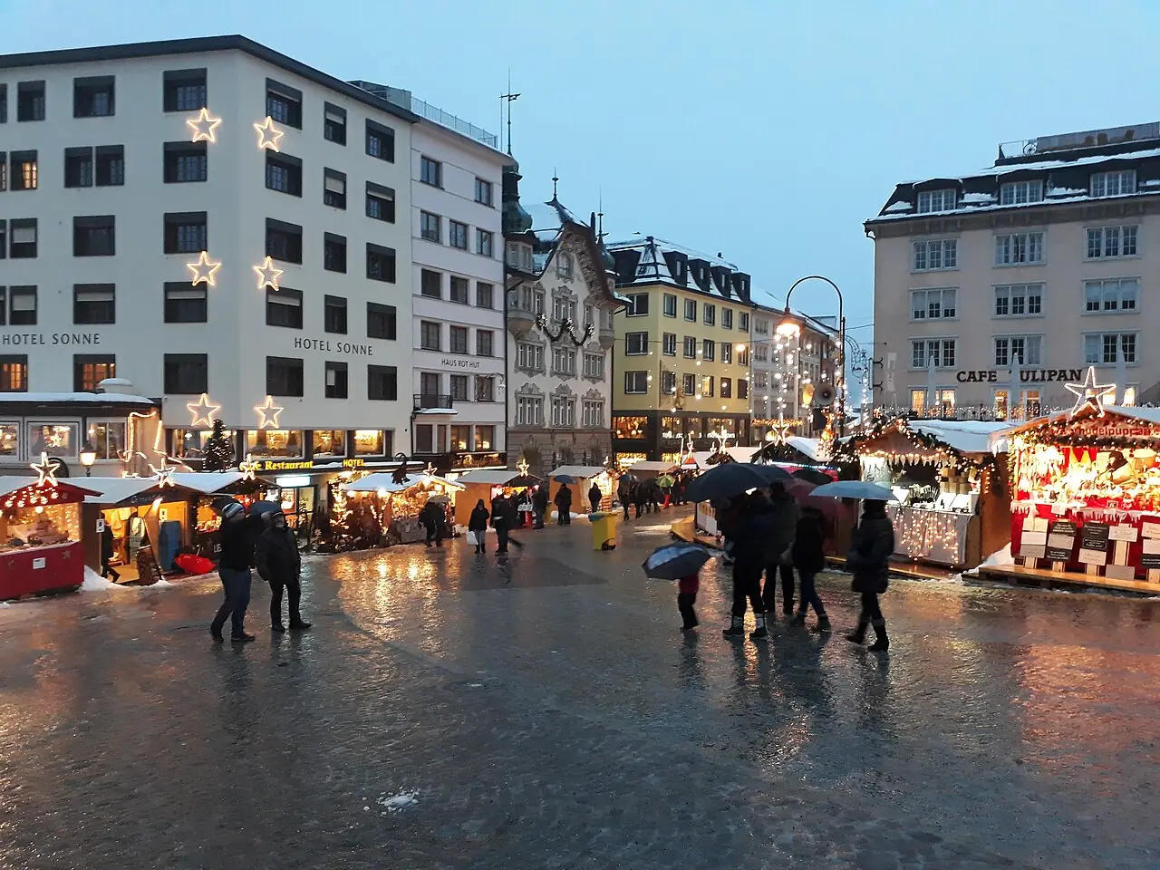 Illuminated market huts on the Hauptplatz at dusk, wet ground and remnants of snow (2017)