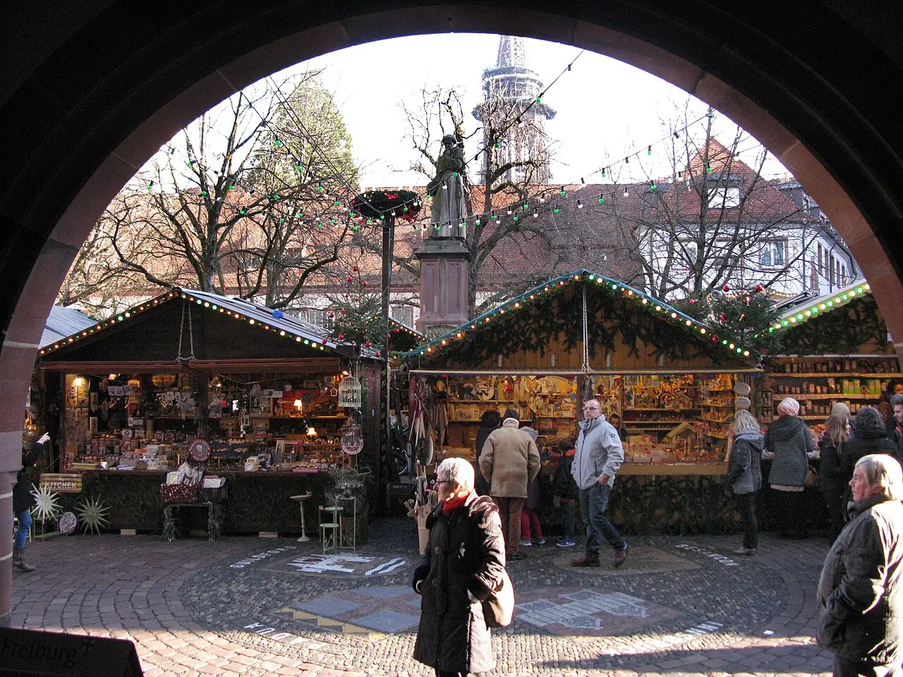 View through an arcade arch onto stalls on Rathausplatz with the Berthold Schwarz monument, the Minster tower in the background, by day