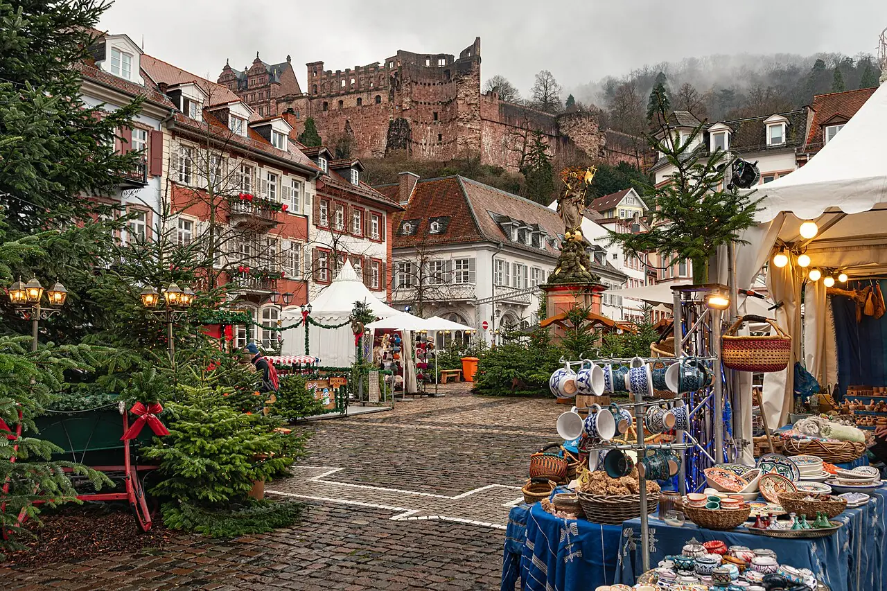 Winter woodland on Kornmarkt with fir trees, white pointed tents and a Madonna statue, the castle above, by day