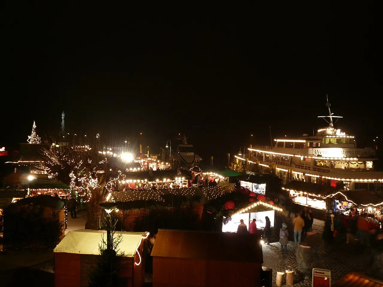 View over the illuminated huts at the harbour at night, a decorated ship on the right, the Imperia statue in the background