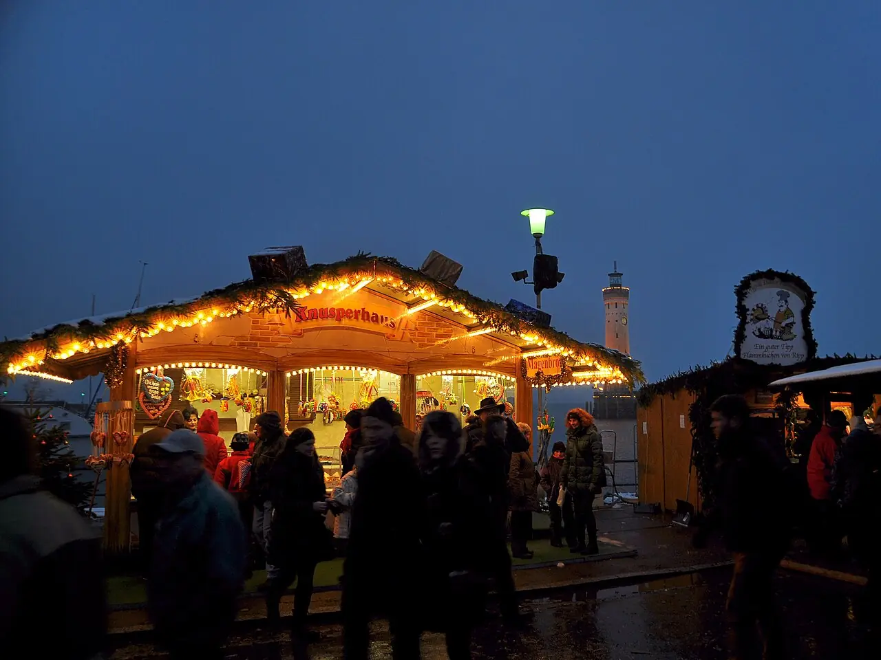 Illuminated hut with gingerbread hearts at blue hour, with the lighthouse at the harbour entrance behind