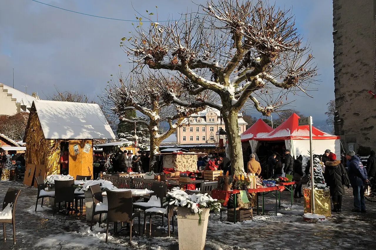Snow-covered huts and tables under plane trees near the Mangturm, with visitors