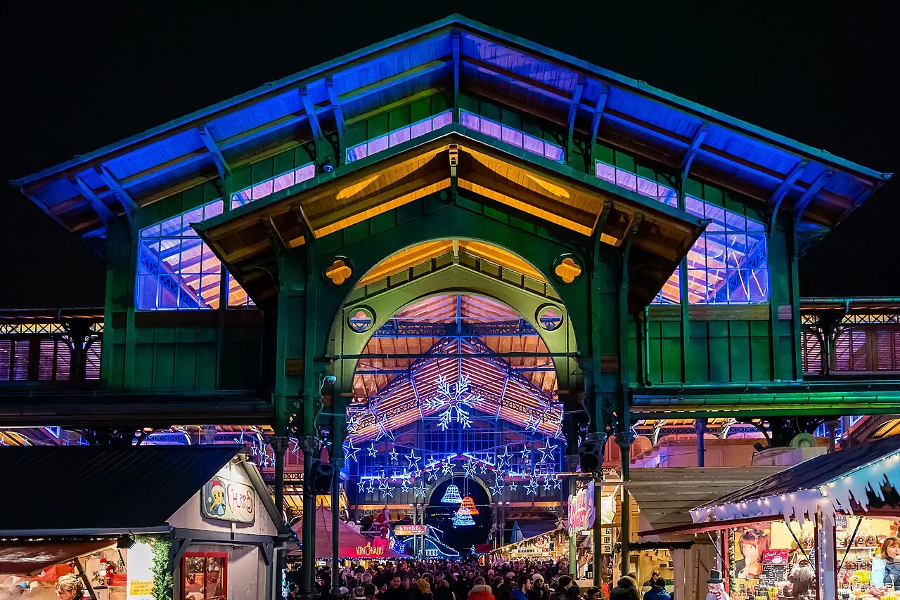Illuminated market hall (Marché couvert) on Place du Marché at night, with chalets and visitors in front (2016)