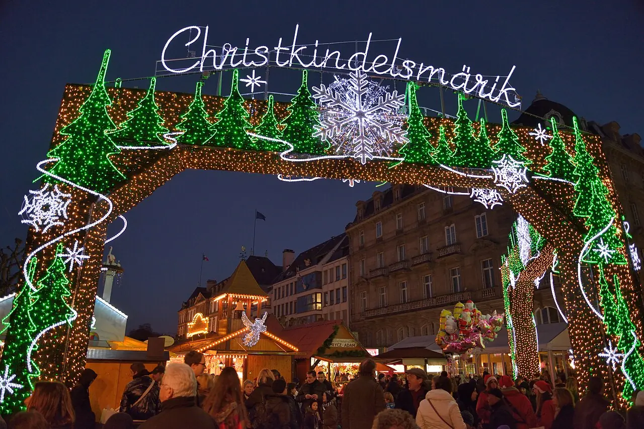 Illuminated gate with the lettering «Christkindelsmärik» at the entrance to Place Broglie, chalets behind it, in the evening